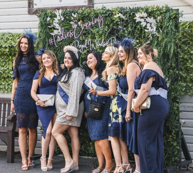 A hen do party in blue and gold poses in front of the flower wall at Windsor Racecourse